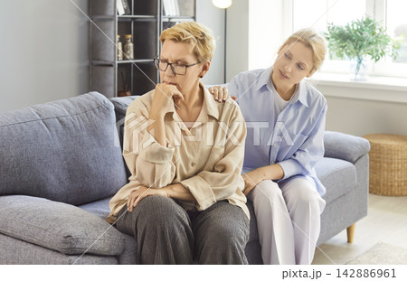 Young woman comforting an elderly female relative on a couch at home gently Young woman comforting an elderly female relative on a couch at home gently 142886961