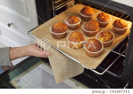 Person carefully removes tray of freshly baked cupcakes with golden brown tops from oven. Person carefully removes tray of freshly baked cupcakes with golden brown tops from oven. 142887341
