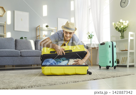 Happy man sitting on floor in living room at home packing suitcases ready for summer vacation Happy man sitting on floor in living room at home packing suitcases ready for summer vacation 142887342