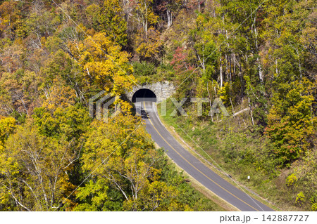 Car driving at mountain road tunnel surrounded by dense woodland in peak autumn. Golden, orange and crimson leaves create breathtaking landscape. Car driving at mountain road tunnel surrounded by dense woodland in peak autumn. Golden, orange and crimson leaves create breathtaking landscape. 142887727