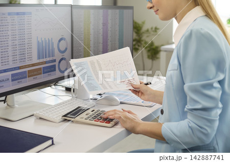 Professional woman accountant reviewing charts using calculator at dual monitors 142887741