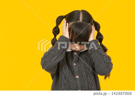 Sad little girl covering her face with hands while standing against a solid background. 142887808