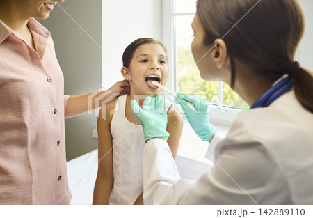 Pediatrician checks child throat with tongue depressor as mother nearby in clinic Pediatrician checks child throat with tongue depressor as mother nearby in clinic 142889110