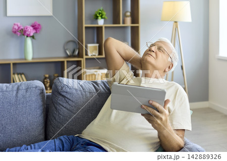 Elderly man with tablet on couch at home, stretching neck and relaxing indoors 142889326
