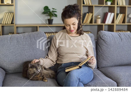 Calm woman reading a book beside her cat on sofa at home, relaxing cozy mood Calm woman reading a book beside her cat on sofa at home, relaxing cozy mood 142889484