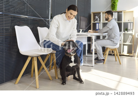 Woman pet owner with dog waiting at veterinary clinic while male vet works at desk Woman pet owner with dog waiting at veterinary clinic while male vet works at desk 142889493