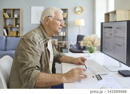 Elderly man manages utility bills on computer with paperwork and calculator at home 142889866