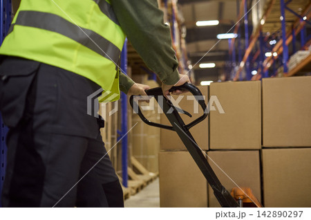 Closeup of warehouse worker steering pallet jack toward boxes on shelves Closeup of warehouse worker steering pallet jack toward boxes on shelves 142890297