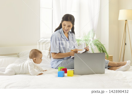 Mother working on laptop with baby crawling among colorful blocks on bed at home Mother working on laptop with baby crawling among colorful blocks on bed at home 142890432