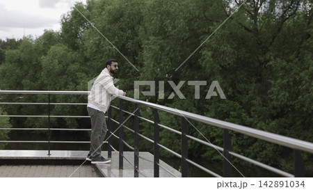 Man leaning on stainless steel railing at riverside viewing platform, gazing toward tree lined river beside park greenery beneath soft overcast daylight, with man, leaning, railing, beard detail Man leaning on stainless steel railing at riverside viewing platform, gazing toward tree lined river beside park greenery beneath soft overcast daylight, with man, leaning, railing, beard detail 142891034