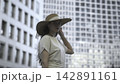 Woman in a wide hat standing before blurred city buildings 142891161