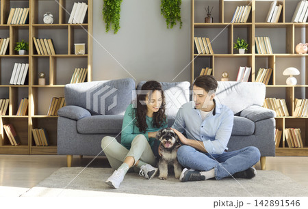 Happy couple sitting on floor at home cuddling mini schnauzer dog in livingroom 142891546