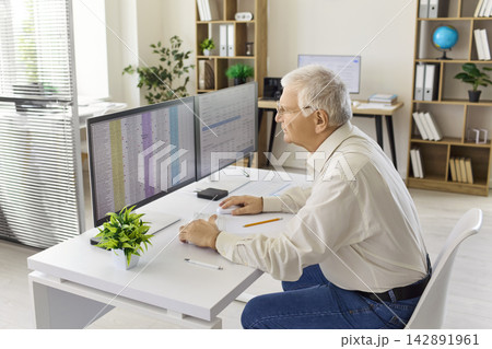 Senior accountant analyzing finance data on dual monitors at modern office desk 142891961