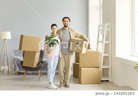 Happy couple carrying box and plant during home move, unpacking in new apartment Happy couple carrying box and plant during home move, unpacking in new apartment 142892263
