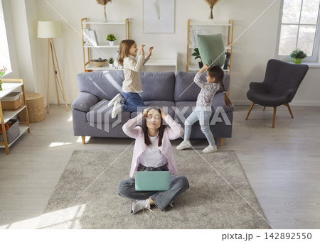 Mother working from home with naughty kids playing pillow fight game by couch Mother working from home with naughty kids playing pillow fight game by couch 142892550