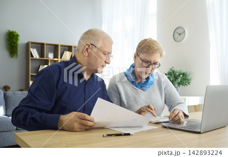 Elderly couple managing finances at home with laptop, review bills and paperwork 142893224