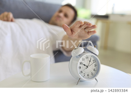 Sleepy man reaching to alarm clock in bed, morning wake-up with coffee mug Sleepy man reaching to alarm clock in bed, morning wake-up with coffee mug 142893384