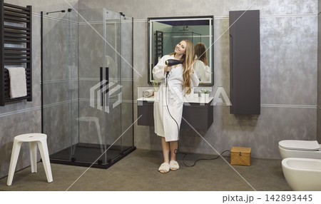 Beautiful young girl drying hair using hairdryer standing in modern bathroom interior 142893445