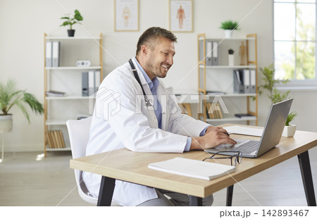 Happy doctor sitting at the desk and working in office on a laptop computer. Happy doctor sitting at the desk and working in office on a laptop computer. 142893467