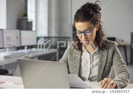 Focused female accountant analyzing financial report with laptop in modern office 142893557