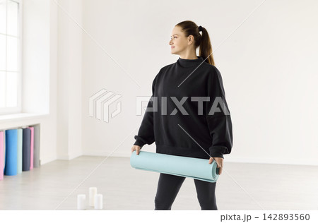 Woman in bright studio holding rolled yoga mat before wellness practice session 142893560