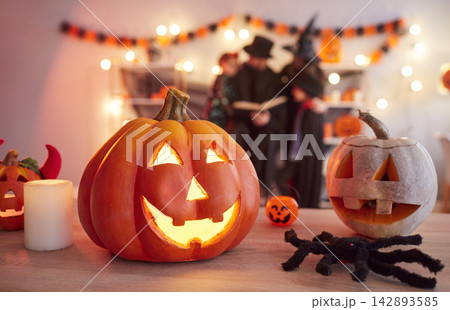 Glowing carved pumpkins standing on table while family in Halloween costumes reading book together. 142893585