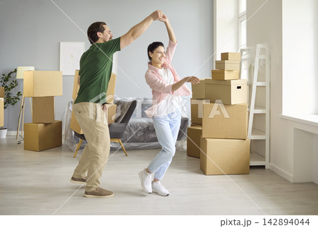 Happy couple dancing among moving boxes in bright new apartment, celebrating home Happy couple dancing among moving boxes in bright new apartment, celebrating home 142894044