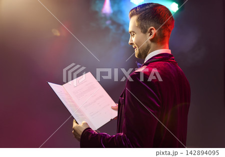 Confident male presenter reading event script on stage under colorful lights 142894095
