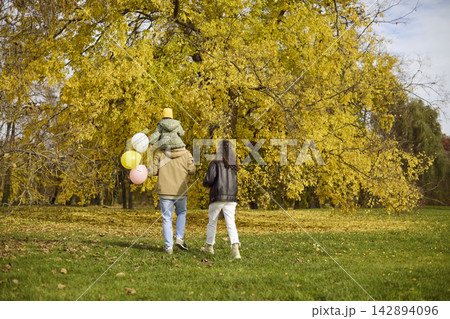 Happy family walking in autumn park with daughter and balloons under golden trees Happy family walking in autumn park with daughter and balloons under golden trees 142894096