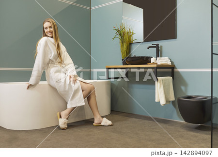 Pretty young woman sitting on bathtub wearing white bathrobe relaxing in modern bathroom interior 142894097