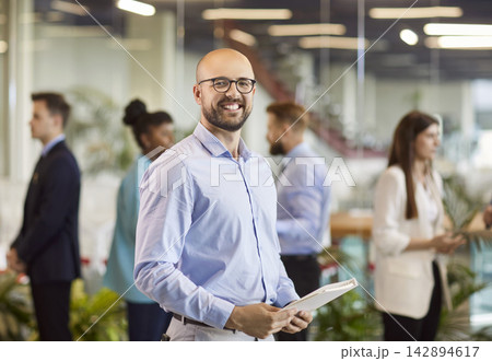 Portrait of smiling bald man wearing glasses looking at camera with company employees on background. 142894617