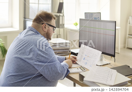Overworked accountant man analyzing budget paperwork at computer in bright office Overworked accountant man analyzing budget paperwork at computer in bright office 142894704