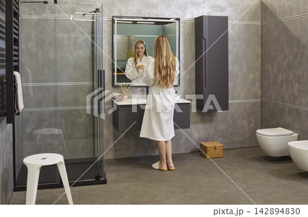 Young cheerful pretty woman standing back in front of mirror in modern bathroom 142894830