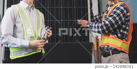 Two Asian engineers wearing hardhats and safety vests discussing solar panel installation and renewable energy project in the office. Two Asian engineers wearing hardhats and safety vests discussing solar panel installation and renewable energy project in the office. 142896161