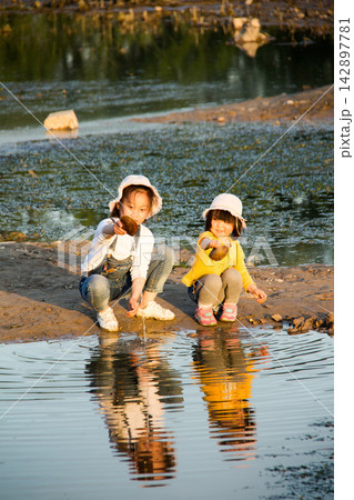 Two little sisters playing with water and reflection by the riverbank Two little sisters playing with water and reflection by the riverbank 142897781