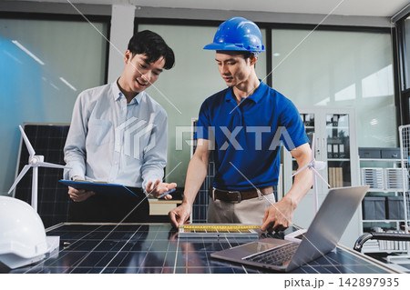Two Asian engineers wearing hardhats and safety vests discussing solar panel installation and renewable energy project in the office. Two Asian engineers wearing hardhats and safety vests discussing solar panel installation and renewable energy project in the office. 142897935