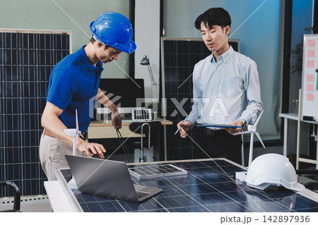 Two Asian engineers wearing hardhats and safety vests discussing solar panel installation and renewable energy project in the office. Two Asian engineers wearing hardhats and safety vests discussing solar panel installation and renewable energy project in the office. 142897936
