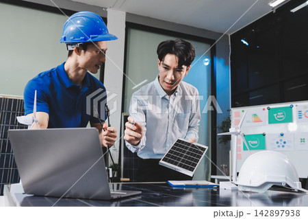 Two Asian engineers wearing hardhats and safety vests discussing solar panel installation and renewable energy project in the office. Two Asian engineers wearing hardhats and safety vests discussing solar panel installation and renewable energy project in the office. 142897938