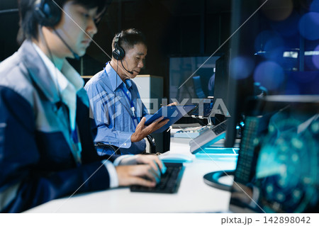 Asian male IT specialist with headset typing on keyboard, monitoring futuristic cybersecurity dashboard and system analytics on computer screen in command center. 142898042