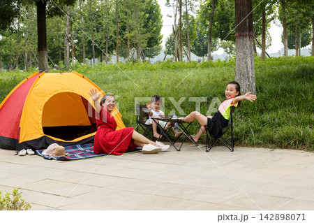 Young family enjoying summer camping in city park with orange tent under green trees Young family enjoying summer camping in city park with orange tent under green trees 142898071