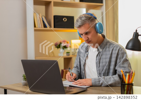 Middle-aged man on home table learning from online course on laptop pausing video and writing notes 142898352