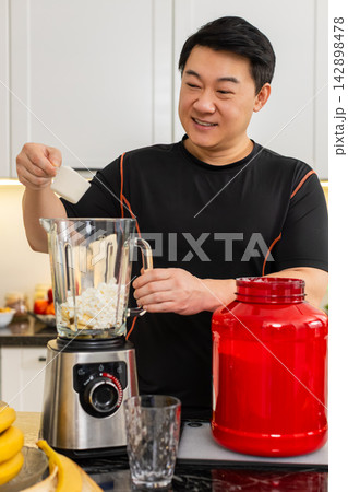 Asian man pouring whey protein into blender in kitchen preparing banana cottage cheese protein shake 142898478