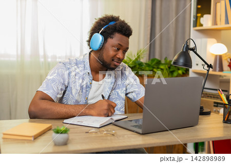 African American young man on home table taking online course listening and writing notes for study African American young man on home table taking online course listening and writing notes for study 142898989