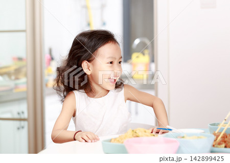 Charming little girl sitting at table during mealtime, looking at camera with a sweet smile, home dining concept. 142899425