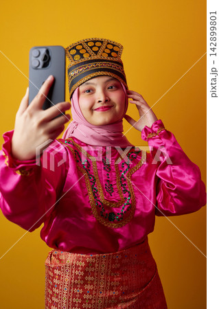 Young Indonesian Asian Woman Taking Selfie in Bright Traditional Dress and Embroidered Headpiece With Hijab 142899801