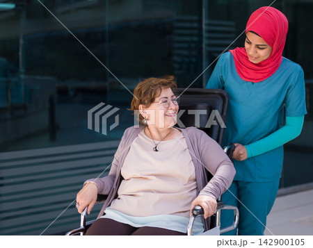 Middle Eastern nurse assisting elderly patient in wheelchair at hospital clinic with compassionate care Middle Eastern nurse assisting elderly patient in wheelchair at hospital clinic with compassionate care 142900105