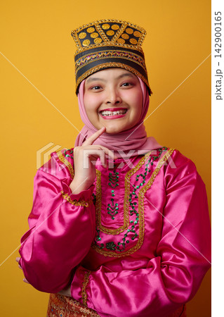 Smiling Young Indonesian Asian Woman In Pink Traditional Costume And Hijab Wearing Ornate Gold Headpiece On Yellow Smiling Young Indonesian Asian Woman In Pink Traditional Costume And Hijab Wearing Ornate Gold Headpiece On Yellow 142900165