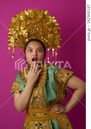 Surprised Young Indonesian Asian Girl Wearing Gold Headdress and Traditional Costume Against Pink Background Surprised Young Indonesian Asian Girl Wearing Gold Headdress and Traditional Costume Against Pink Background 142900187