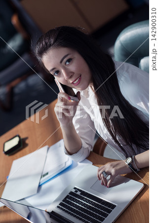 Female business professional on phone working at laptop in a modern open office during a meeting 142900310