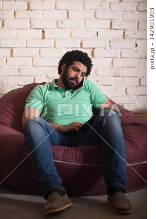Young Arab entrepreneur in a coworking space talking on phone and working on tablet in relaxed startup office 142901903